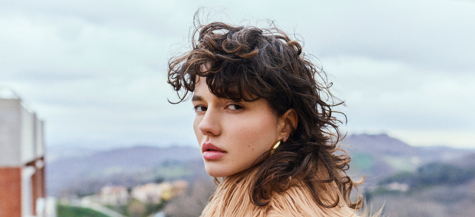 Woman with curly hair wearing a beige fur coat outdoors with a scenic background